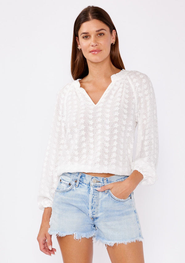 [Color: White] A brunette model wearing a pretty floral embroidered top with eyelet details. A summer breezy top with a ruffled split v neckline and long sleeves with an elastic cuff. An everyday white top styled with cut off denim  shorts for summer. 