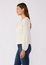 [Color: Ivory] A brunette model wearing a n ivory off white shimmer blouse crafted on textured chiffon. With a split v neckline with ties, long sleeves, and an elastic hem. The perfect special occasion blouse for formal events or holidays