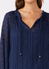 [Color: Navy] A brunette model wearing a navy shimmer blouse crafted on textured chiffon. With a split v neckline with ties, long sleeves, and an elastic hem. The perfect special occasion blouse for formal events or holidays.   