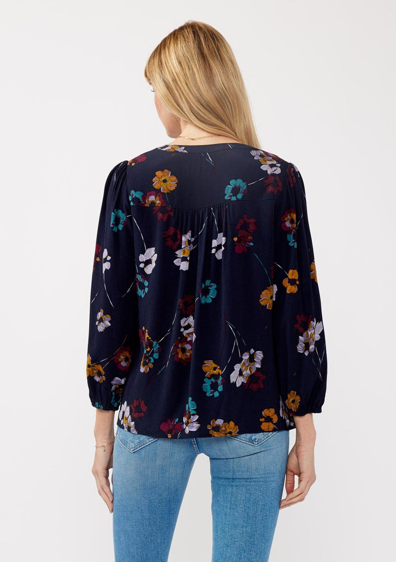 [Color: Navy/Mustard] A back facing image of a blonde model wearing a bohemian fall blouse in a blue floral print. With long sleeves, a v neckline, a self covered button front, and a relaxed fit. 