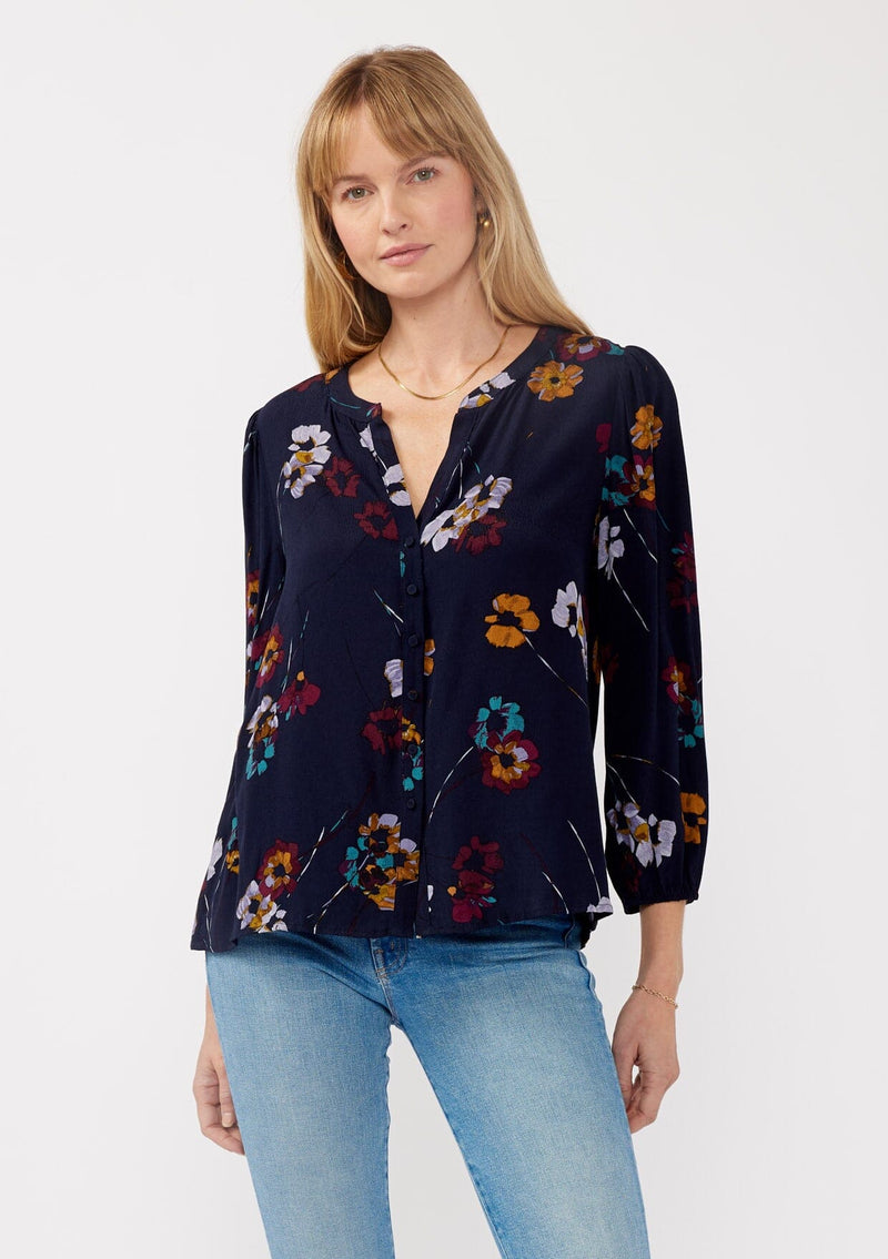 [Color: Navy/Mustard] A front facing image of a blonde model wearing a bohemian fall blouse in a blue floral print. With long sleeves, a v neckline, a self covered button front, and a relaxed fit. 