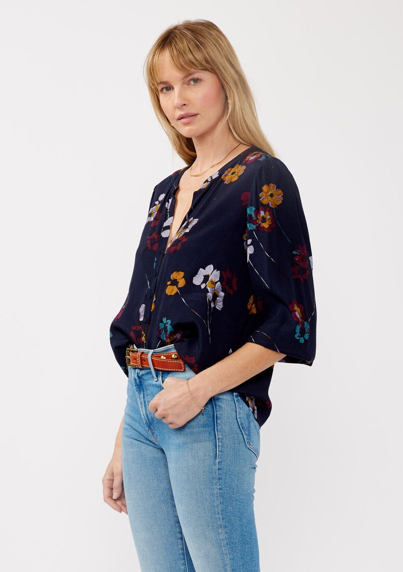 [Color: Navy/Mustard] A side facing image of a blonde model wearing a bohemian fall blouse in a blue floral print. With long sleeves, a v neckline, a self covered button front, and a relaxed fit. 