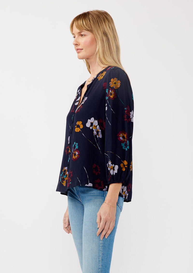 [Color: Navy/Mustard] A side facing image of a blonde model wearing a bohemian fall blouse in a blue floral print. With long sleeves, a v neckline, a self covered button front, and a relaxed fit. 