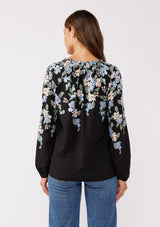 [Color: Black/Pale Blue] A back facing image of brunette model wearing a black and blue floral print top. This relaxed fit top features a smocked split v-neckline, long sleeves, and elastic cuffs. Paired beautifully with denim to compliment the Fall season.  