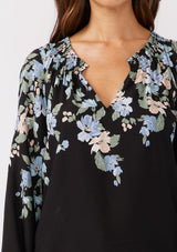 [Color: Black/Pale Blue] A detail shot of brunette model wearing a black and blue floral print top. This relaxed fit top features a smocked split v-neckline, long sleeves, and elastic cuffs. Paired beautifully with denim to compliment the Fall season.  