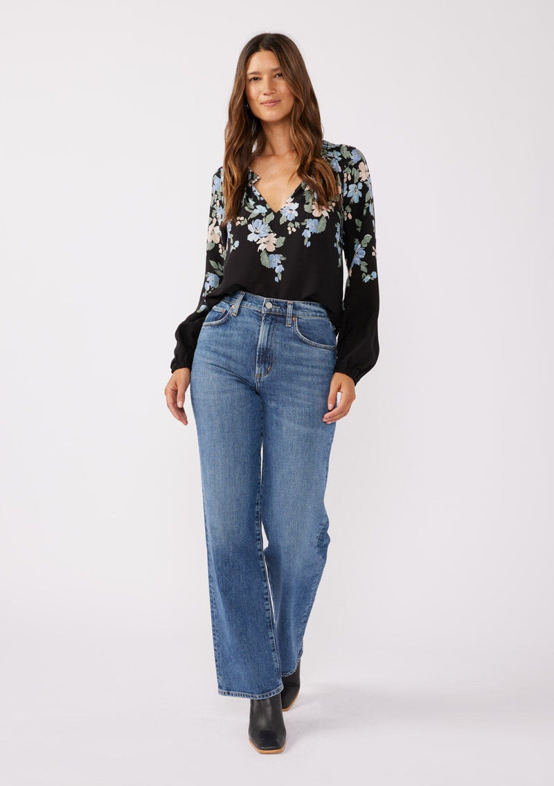 [Color: Black/Pale Blue] A full body front facing image of brunette model wearing a black and blue floral print top. This relaxed fit top features a smocked split v-neckline, long sleeves, and elastic cuffs. Paired beautifully tucked into denim to compliment the Fall season.  