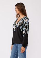 [Color: Black/Pale Blue] A side facing image of brunette model wearing a black and blue floral print top. This relaxed fit top features a smocked split v-neckline, long sleeves, and elastic cuffs. Paired beautifully with denim to compliment the Fall season.  