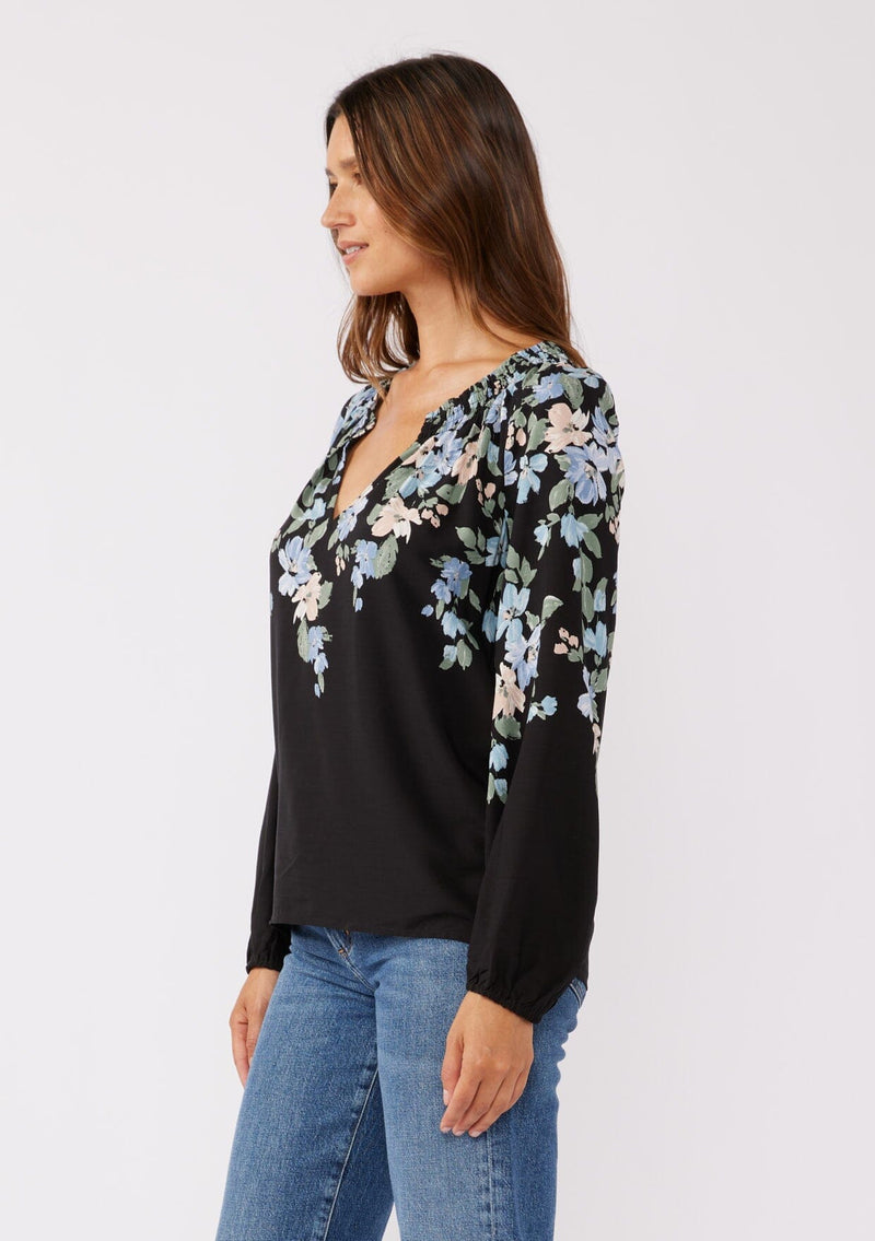 [Color: Black/Pale Blue] A side facing image of brunette model wearing a black and blue floral print top. This relaxed fit top features a smocked split v-neckline, long sleeves, and elastic cuffs. Paired beautifully with denim to compliment the Fall season.  