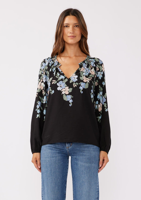 [Color: Black/Pale Blue] A front facing image of brunette model wearing a black and blue floral print top. This relaxed fit top features a smocked split v-neckline, long sleeves, and elastic cuffs. Paired beautifully with denim to compliment the Fall season.  