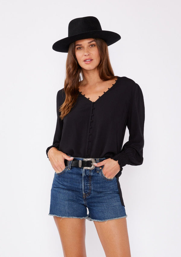 [Color: Black] A front facing image of a brunette model wearing a bohemian black blouse. With long sleeves, side vents, smocked ruffled wrist cuffs, a button loop trimmed v neckline, and a self covered button front.