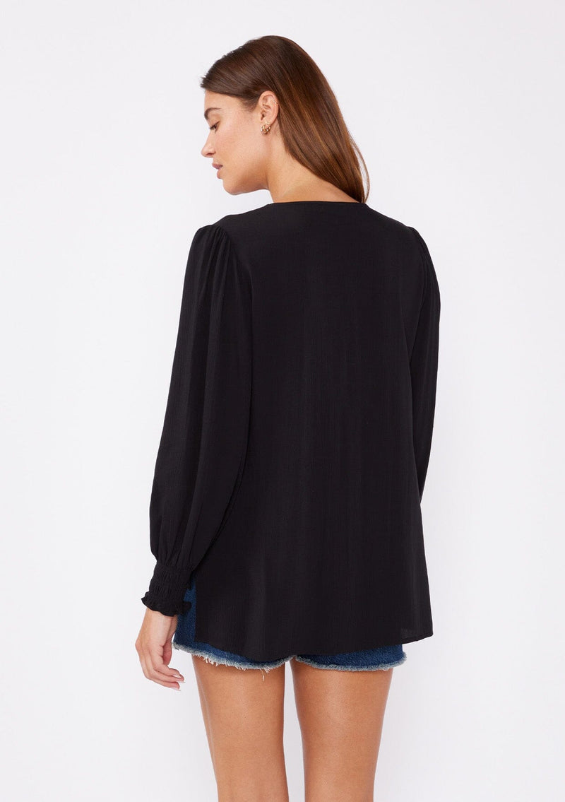 [Color: Black] A back facing image of a brunette model wearing a bohemian black blouse. With long sleeves, side vents, smocked ruffled wrist cuffs, a button loop trimmed v neckline, and a self covered button front.