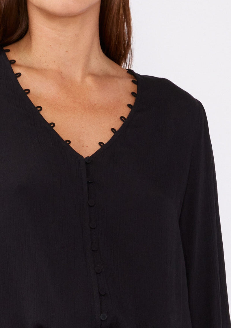 [Color: Black] A detail mage of a brunette model wearing a bohemian black blouse. With long sleeves, side vents, smocked ruffled wrist cuffs, a button loop trimmed v neckline, and a self covered button front.