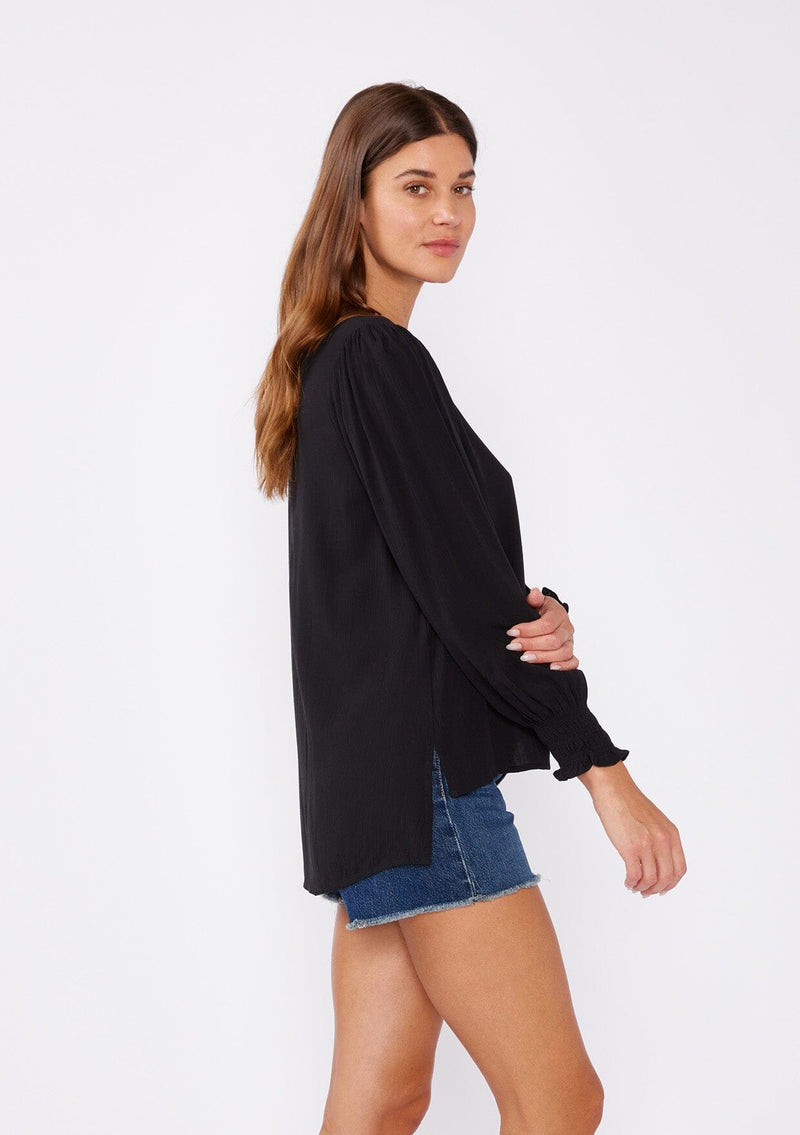 [Color: Black] A side facing image of a brunette model wearing a bohemian black blouse. With long sleeves, side vents, smocked ruffled wrist cuffs, a button loop trimmed v neckline, and a self covered button front.