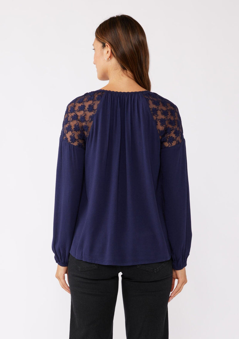 [Color: Navy] A back facing image of a brunette model wearing a navy blue bohemian fall blouse with long sleeves, a split v neckline with ties, and sheer embroidered mesh detail along the shoulders.