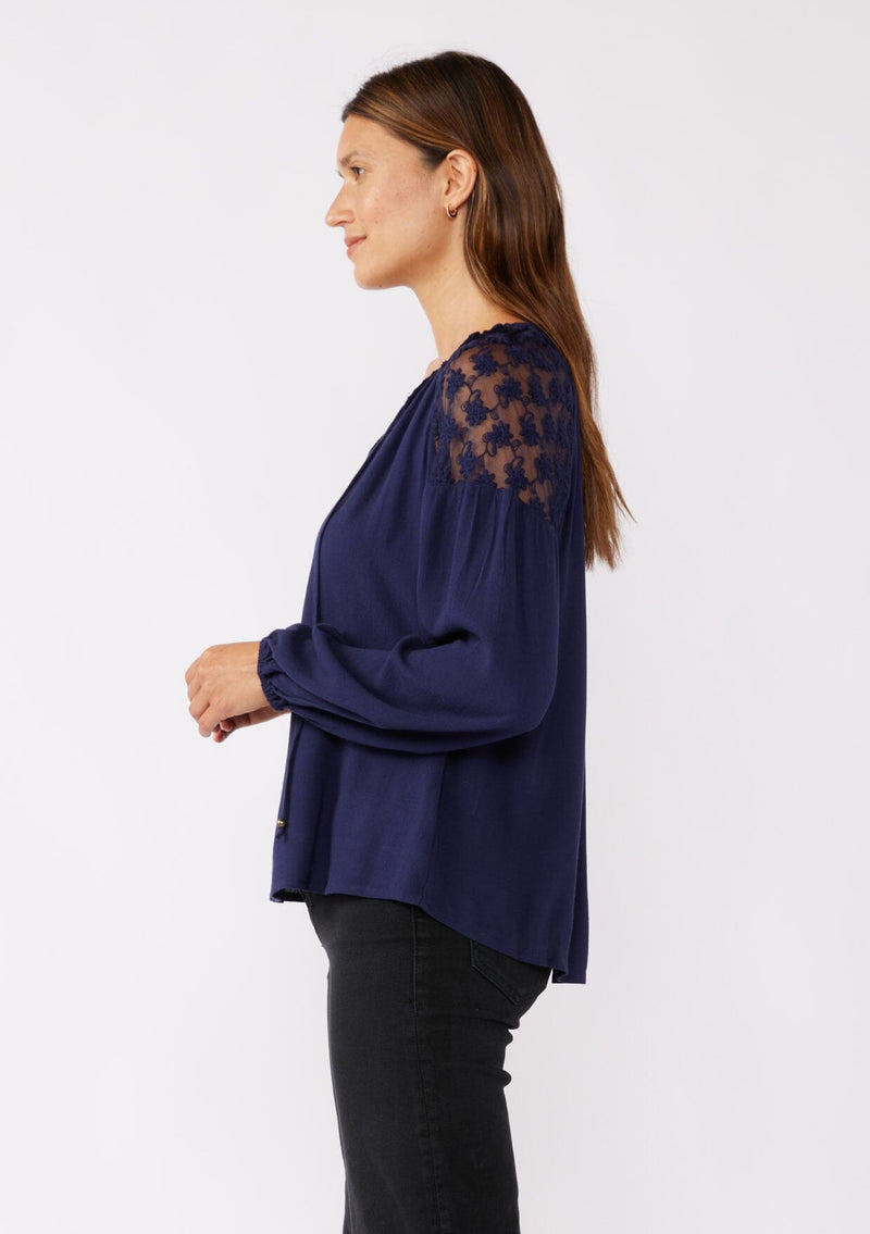 [Color: Navy] A side facing image of a brunette model wearing a navy blue bohemian fall blouse with long sleeves, a split v neckline with ties, and sheer embroidered mesh detail along the shoulders.