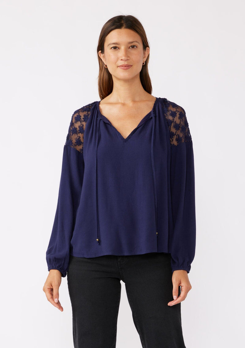 [Color: Navy] A front facing image of a brunette model wearing a navy blue bohemian fall blouse with long sleeves, a split v neckline with ties, and sheer embroidered mesh detail along the shoulders.