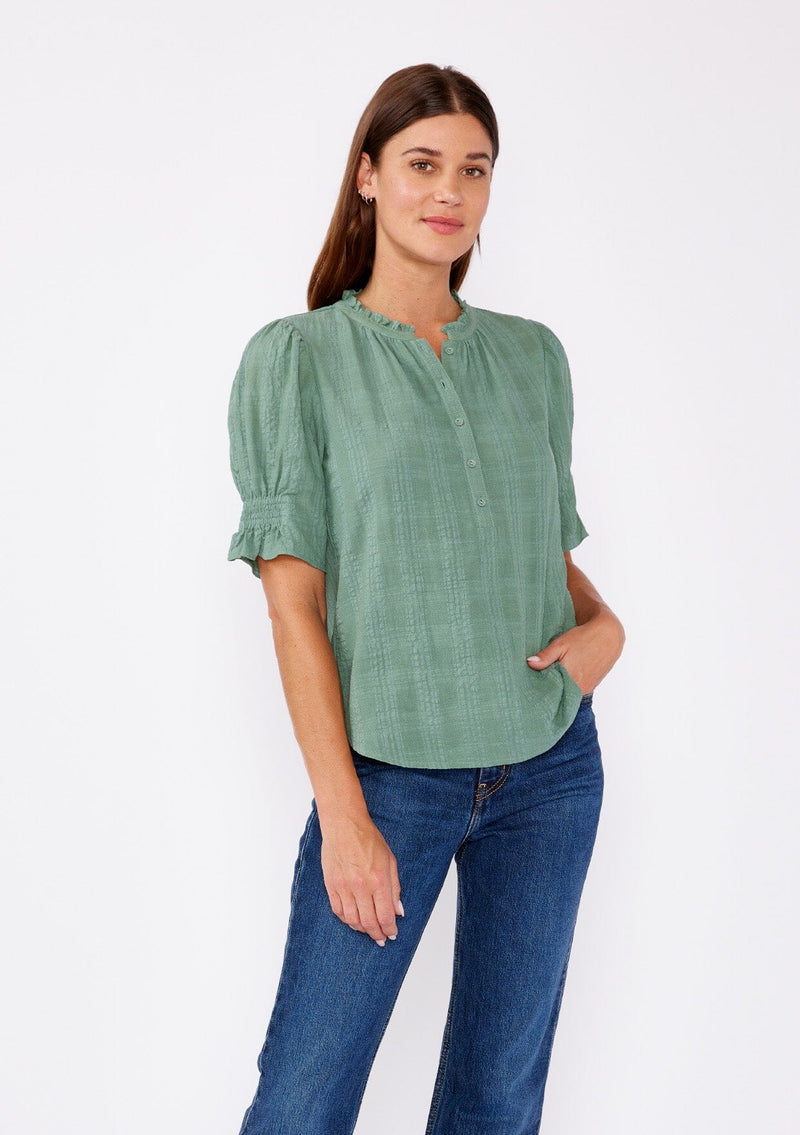 [Color: Dusty Olive] A front facing image of a blonde model wearing a green bohemian cotton blend blouse in a textured gingham. With short puff sleeves, a button front, a high ruffled neckline, and smocked elastic details at the sleeve.