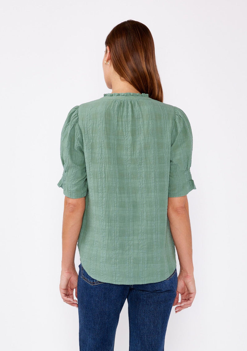 [Color: Dusty Olive] A back facing image of a blonde model wearing a green bohemian cotton blend blouse in a textured gingham. With short puff sleeves, a button front, a high ruffled neckline, and smocked elastic details at the sleeve.