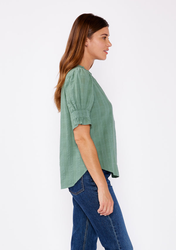 [Color: Dusty Olive] A side facing image of a blonde model wearing a green bohemian cotton blend blouse in a textured gingham. With short puff sleeves, a button front, a high ruffled neckline, and smocked elastic details at the sleeve.