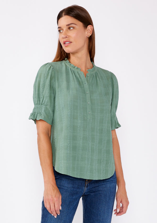 [Color: Dusty Olive] A front facing image of a blonde model wearing a green bohemian cotton blend blouse in a textured gingham. With short puff sleeves, a button front, a high ruffled neckline, and smocked elastic details at the sleeve.