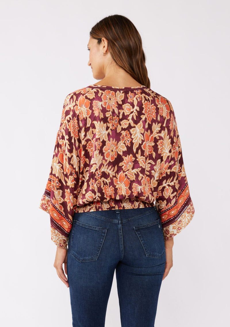 [Color: Wine/Dusty Rose] A back image of a brunette model wearing an orange and red bohemian floral border print blouse. Perfectly suited for the fall season, with flowy kimono sleeves, button front, flattering v neckline, and half smocked elastic waist at the back. A trendy and stylish boho top paired with vintage denim. 