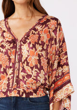 [Color: Wine/Dusty Rose] A detailed image of a brunette model wearing an orange and red bohemian floral border print blouse. Perfectly suited for the fall season, with flowy kimono sleeves, button front, flattering v neckline, and half smocked elastic waist at the back. A trendy and stylish boho top paired with vintage denim. 