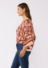 [Color: Wine/Dusty Rose] A side image of a brunette model wearing an orange and red bohemian floral border print blouse. Perfectly suited for the fall season, with flowy kimono sleeves, button front, flattering v neckline, and half smocked elastic waist at the back. A trendy and stylish boho top paired with vintage denim. 