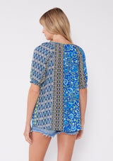 [Color: Blue/Lime] A blonde model wearing an ultra bohemian blouse with a multi print blue floral design. A summer top with a split v neckline, tassel tie front, and short raglan puff sleeves. The perfect summer top for any casual occasion.