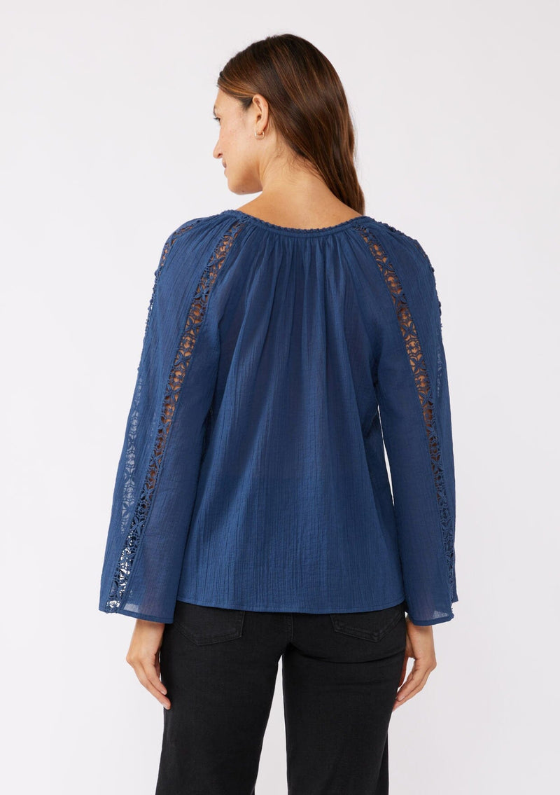 [Color: Blue Indigo] A back facing image of a brunette model wearing a classic bohemian blue cotton blouse. With crochet trim, a split v neckline, and long bell sleeves. 