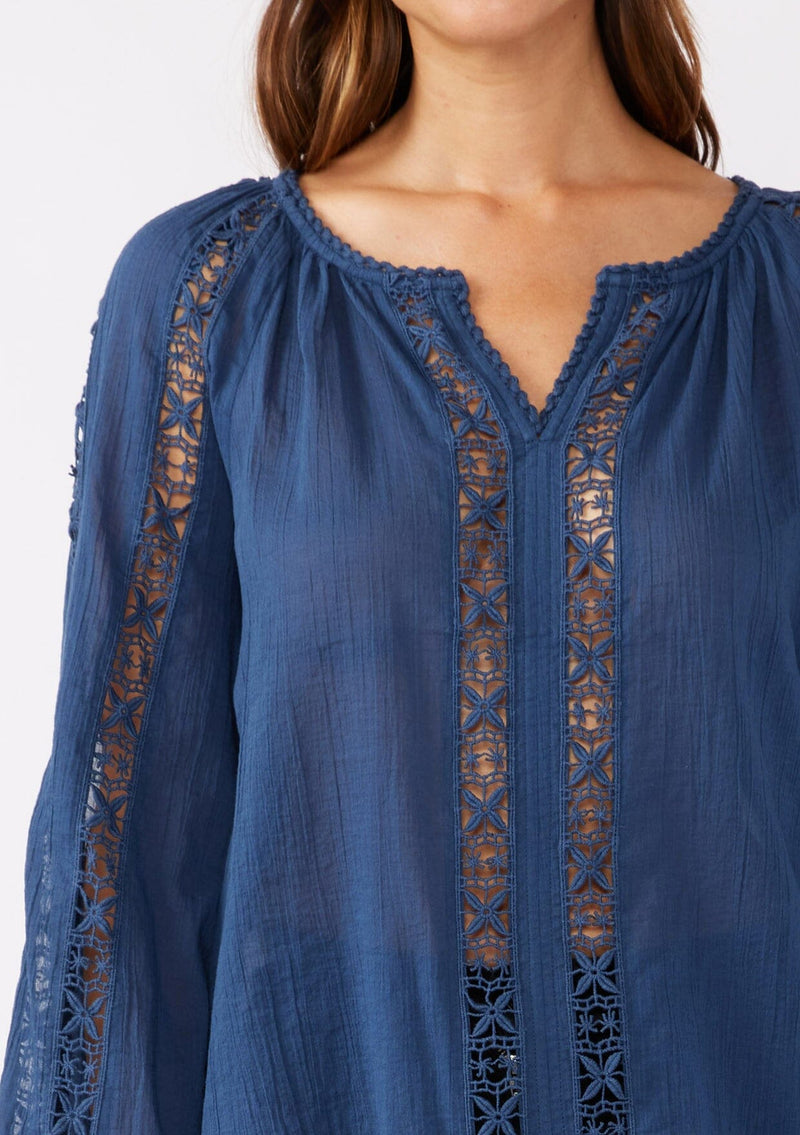 [Color: Blue Indigo] A detailed shot of a brunette model wearing a classic bohemian blue cotton blouse. With crochet trim, a split v neckline, and long bell sleeves. 