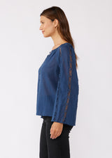 [Color: Blue Indigo] A side facing image of a brunette model wearing a classic bohemian blue cotton blouse. With crochet trim, a split v neckline, and long bell sleeves. 