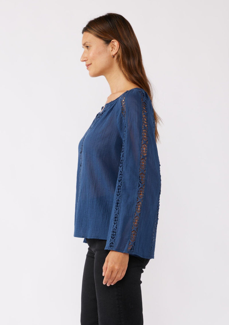 [Color: Blue Indigo] A side facing image of a brunette model wearing a classic bohemian blue cotton blouse. With crochet trim, a split v neckline, and long bell sleeves. 