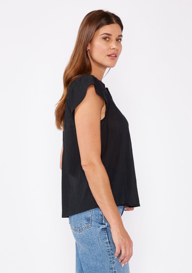 [Color: Black] A blond model wearing a black bohemian top crafted from lightweight cotton gauze. With short cap sleeves, a split v neckline, gathered details at the yoke, and a raw edge hem.