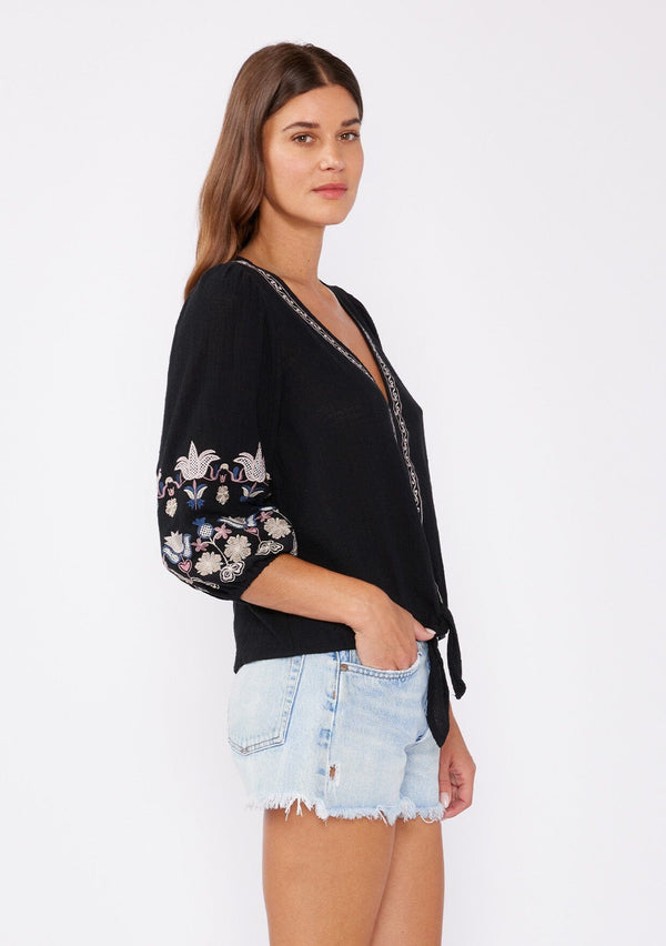 [Color: Black/Dusty Rose] A front facing image of a brunette model wearing an black bohemian blouse with colorful floral embroidered details throughout. Featuring three quarter length sleeves, a v neckline, and a tie waist detail.