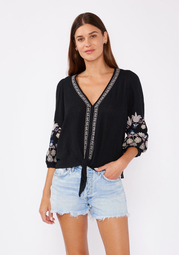 [Color: Black/Dusty Rose] A front facing image of a brunette model wearing an black bohemian blouse with colorful floral embroidered details throughout. Featuring three quarter length sleeves, a v neckline, and a tie waist detail.