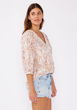 [Color: Cream/Dusty Brick] A brunette model wearing a bohemian cotton blouse in pink ikat print. With a surplice v neckline, long sleeve with button cuff closure, high low elastic hem at front, and crochet trim inserts.