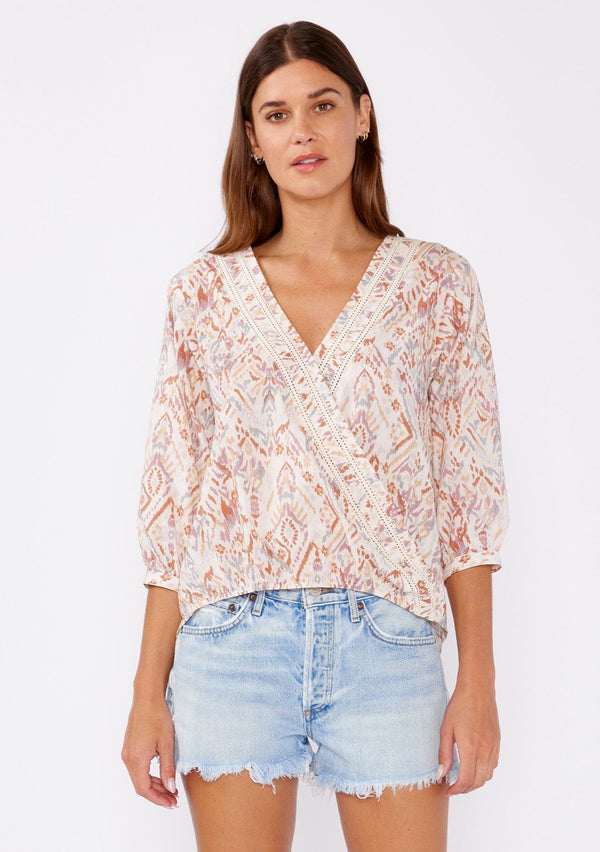 [Color: Cream/Dusty Brick] A brunette model wearing a bohemian cotton blouse in pink ikat print. With a surplice v neckline, long sleeve with button cuff closure, high low elastic hem at front, and crochet trim inserts. 