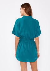 [Color: Forest Teal] A back facing image of a brunette model wearing a teal tunic shirt with a collared neckline, a button front, short sleeves with a dropped shoulder, and a smocked elastic waist.