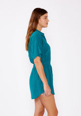 [Color: Forest Teal] A side facing image of a brunette model wearing a teal tunic shirt with a collared neckline, a button front, short sleeves with a dropped shoulder, and a smocked elastic waist.
