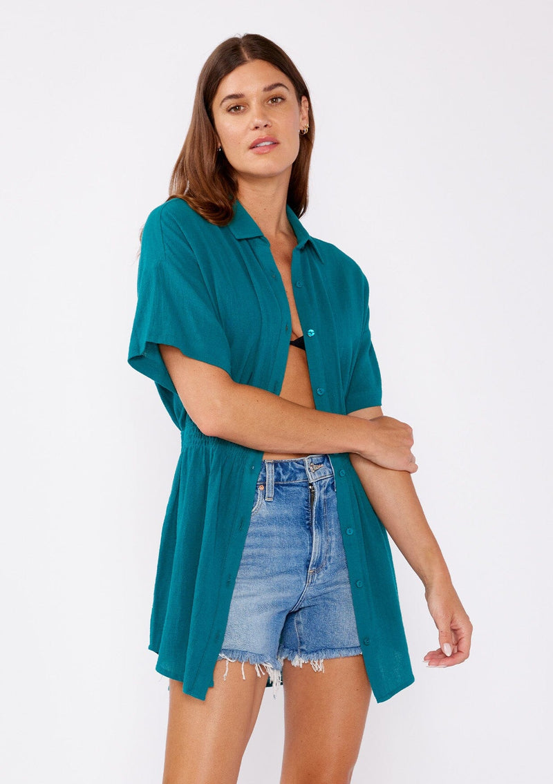 [Color: Forest Teal] A front facing image of a brunette model wearing a teal tunic shirt with a collared neckline, a button front, short sleeves with a dropped shoulder, and a smocked elastic waist.