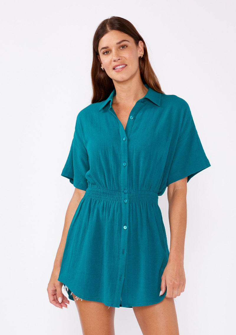 [Color: Forest Teal] A front facing image of a brunette model wearing a teal tunic shirt with a collared neckline, a button front, short sleeves with a dropped shoulder, and a smocked elastic waist.