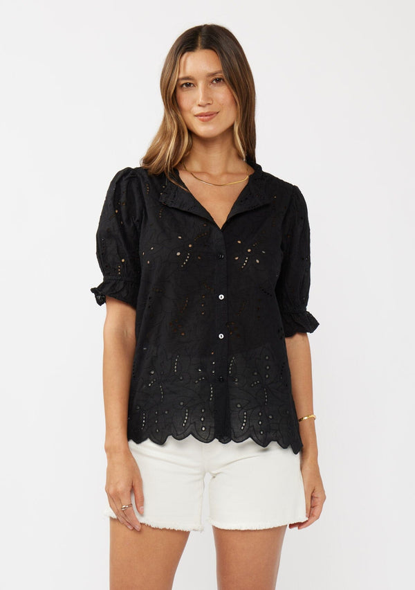 [Color: Black] A front facing image of a brunette model wearing a black eyelet top with short puff sleeves, ruffle trimmed neckline, and button front. 