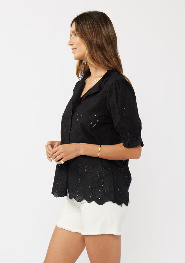 [Color: Black] A side facing image of a brunette model wearing a black eyelet top with short puff sleeves, ruffle trimmed neckline, and button front. 