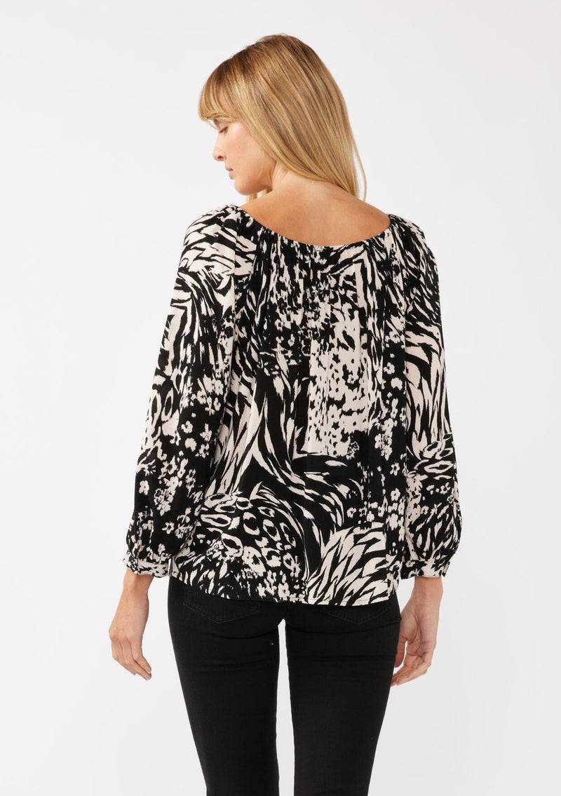 [Color: Black/Blush] A back facing image of a blonde model wearing a bohemian black and white abstract print blouse. Featuring an elastic scoop neckline, tie front detail, long sleeves with cuffs, and a relaxed fit. Perfect for any casual outing whether paired with shorts or long bottoms. 