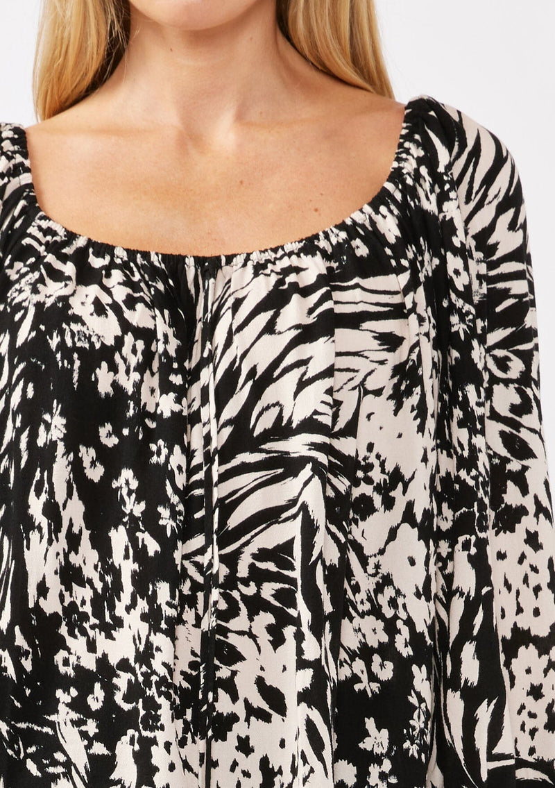 [Color: Black/Blush] A detail image of a blonde model wearing a bohemian black and white abstract print blouse. Featuring an elastic scoop neckline, tie front detail, long sleeves with cuffs, and a relaxed fit. Perfect for any casual outing whether paired with shorts or long bottoms. 