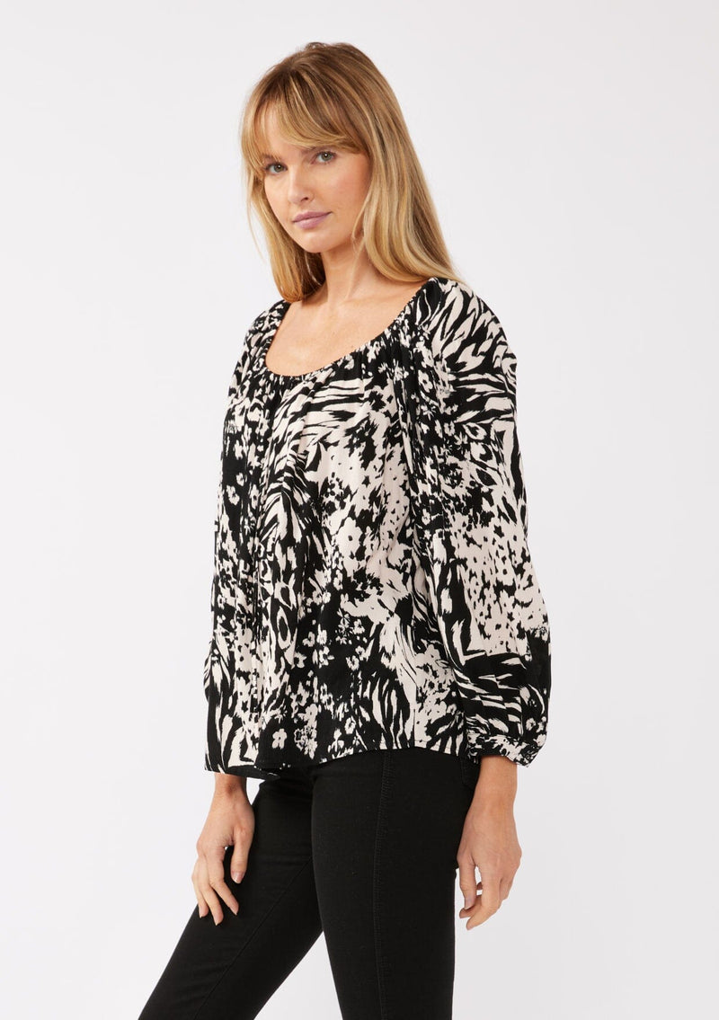 [Color: Black/Blush] A side facing image of a blonde model wearing a bohemian black and white abstract print blouse. Featuring an elastic scoop neckline, tie front detail, long sleeves with cuffs, and a relaxed fit. Perfect for any casual outing whether paired with shorts or long bottoms. 