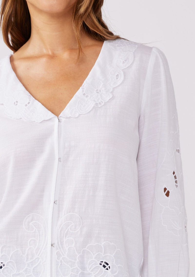 [Color: White] A detailed shot of a brunette model wearing a white blouse with a peter pan collar. A relaxed fit bohemian blouse with tonal embroidery, hook and eye front closure, long sleeves, and ruffle trim cuffs.