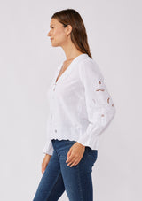 [Color: White] A side facing image of a brunette model wearing a white blouse with a peter pan collar. A relaxed fit bohemian blouse with tonal embroidery, hook and eye front closure, long sleeves, and ruffle trim cuffs.