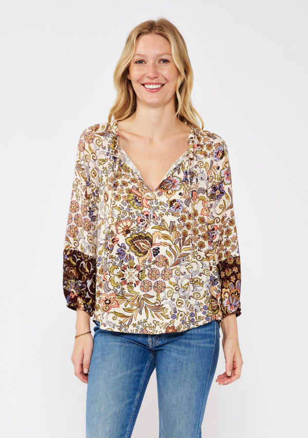 [Color: Natural/Brown] A blonde model wearing a neutral brown top with mixed florals for the fall season. With a split v neckline and tie neck, long sleeve, elastic cuff, and a relaxed fit. The perfect fall top paired with denim jeans.