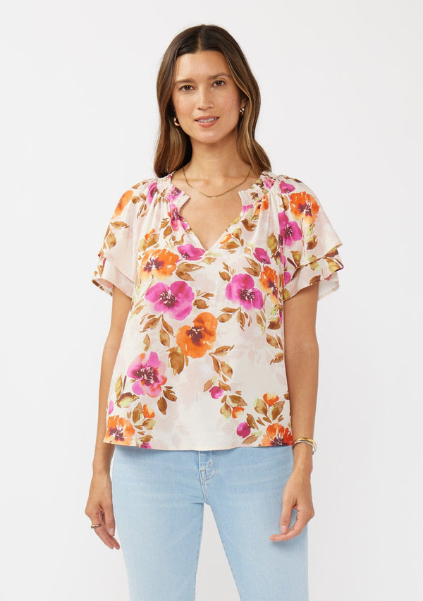 [Color: Natural/Orchid] A front facing image of a brunette model wearing a relaxed fit top in a floral print. With short double flutter sleeves, split v neckline, and a smocked detail at the neck. A dressy but casual floral top styled with light denim.  